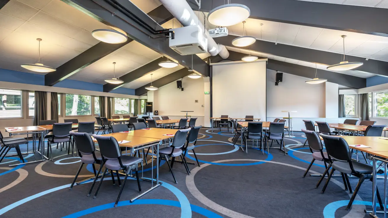 Konferansesal med gruppe bord, store taklamper, prosjektor og lerret og teppebelagt gulv.