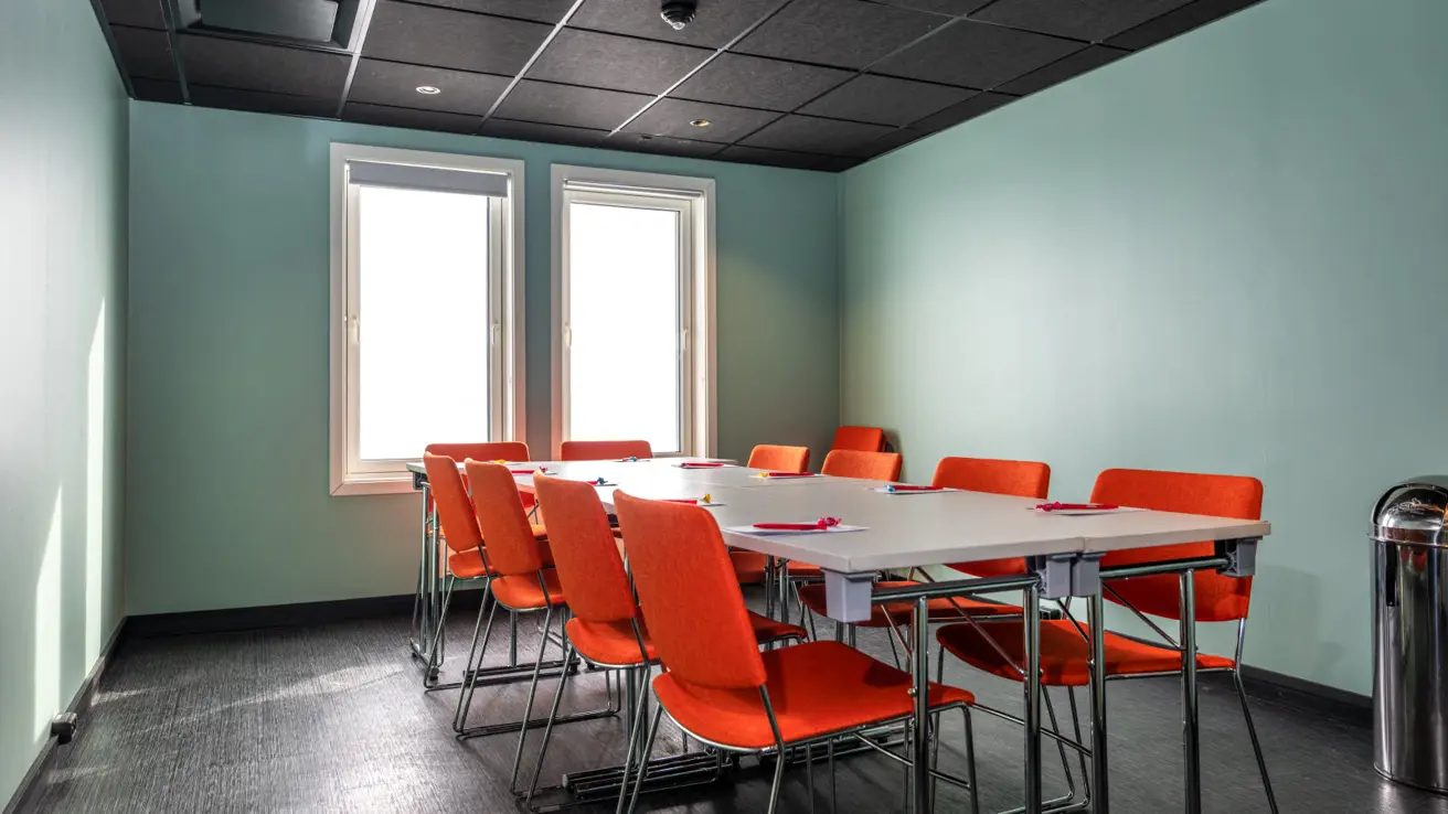 Møterom til ca 10 personer med langbord og oransje stoler rundt. Lys grønn farge på veggen og sort gulv.
