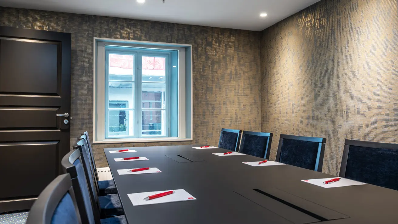 Møterom med langbord og stoler med blå detaljer, tapet på veggene og et vindu i enden av bordet.