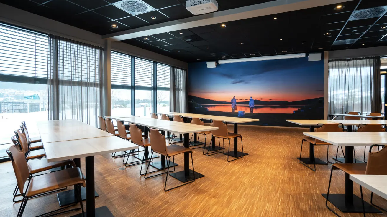 Mor Åse-mødelokale i klasseværelsesopsætning med midtergang mellem siddepladserne. Vinduer langs hele siden + nogle i front. Billede fra Peer Gynt i front. Lærred i front.